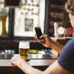 Homem com um celular, bebendo cerveja, enquanto compra cervejas online.